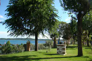 Grüner Campingplatz mit Rasen, Bäumen und einem Blick auf den See, im Hintergrund Ufer und Wasserlinie.
