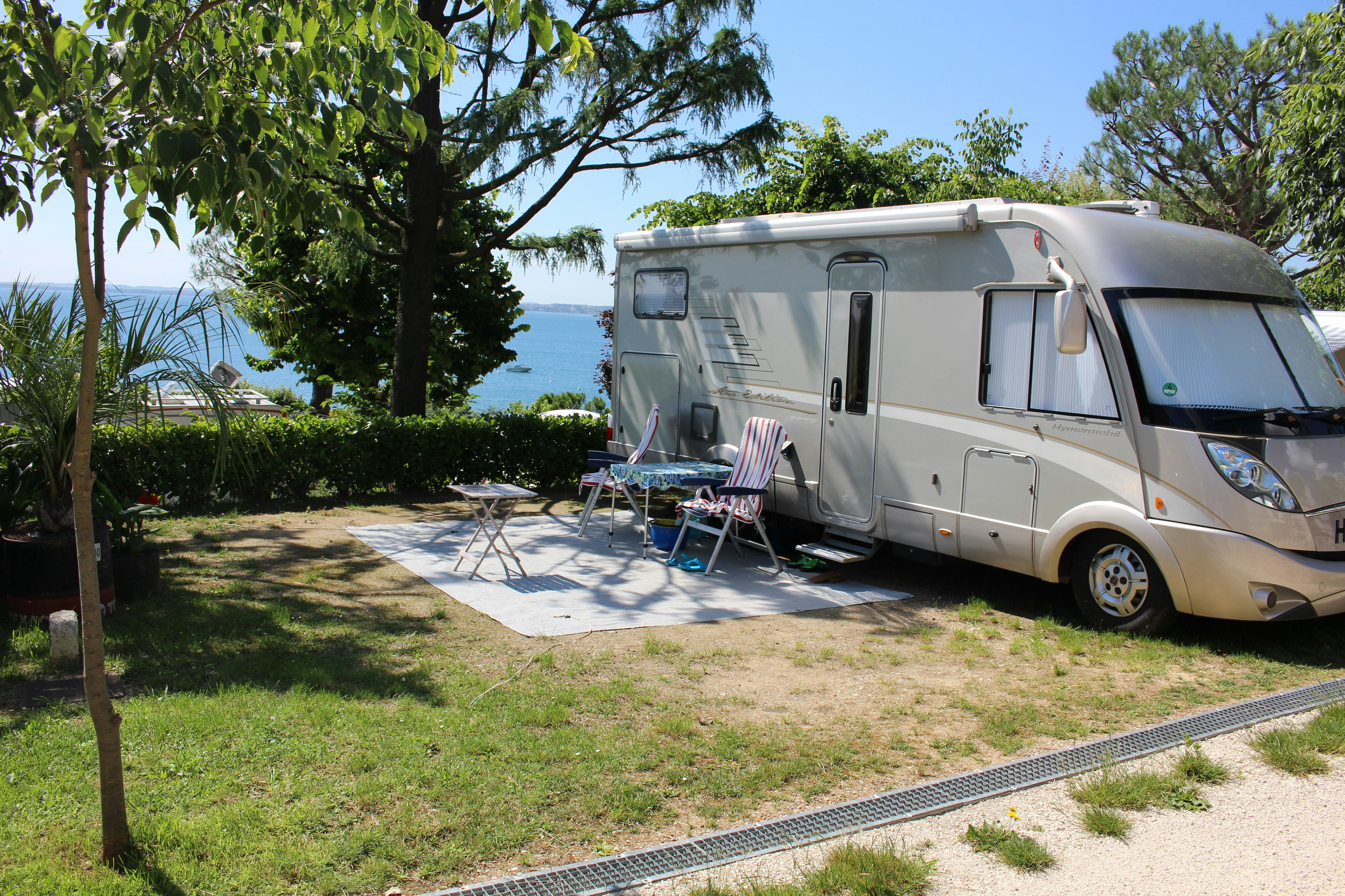 Wohnmobil auf einem Campingplatz mit Sonnenschirm und Stühlen am Stellplatz, Hecken und Meerblick zwischen Bäumen.