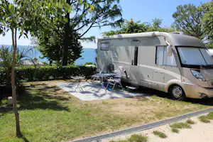 Wohnmobil auf einem Campingplatz mit Sonnenschirm und Stühlen am Stellplatz, Hecken und Meerblick zwischen Bäumen.