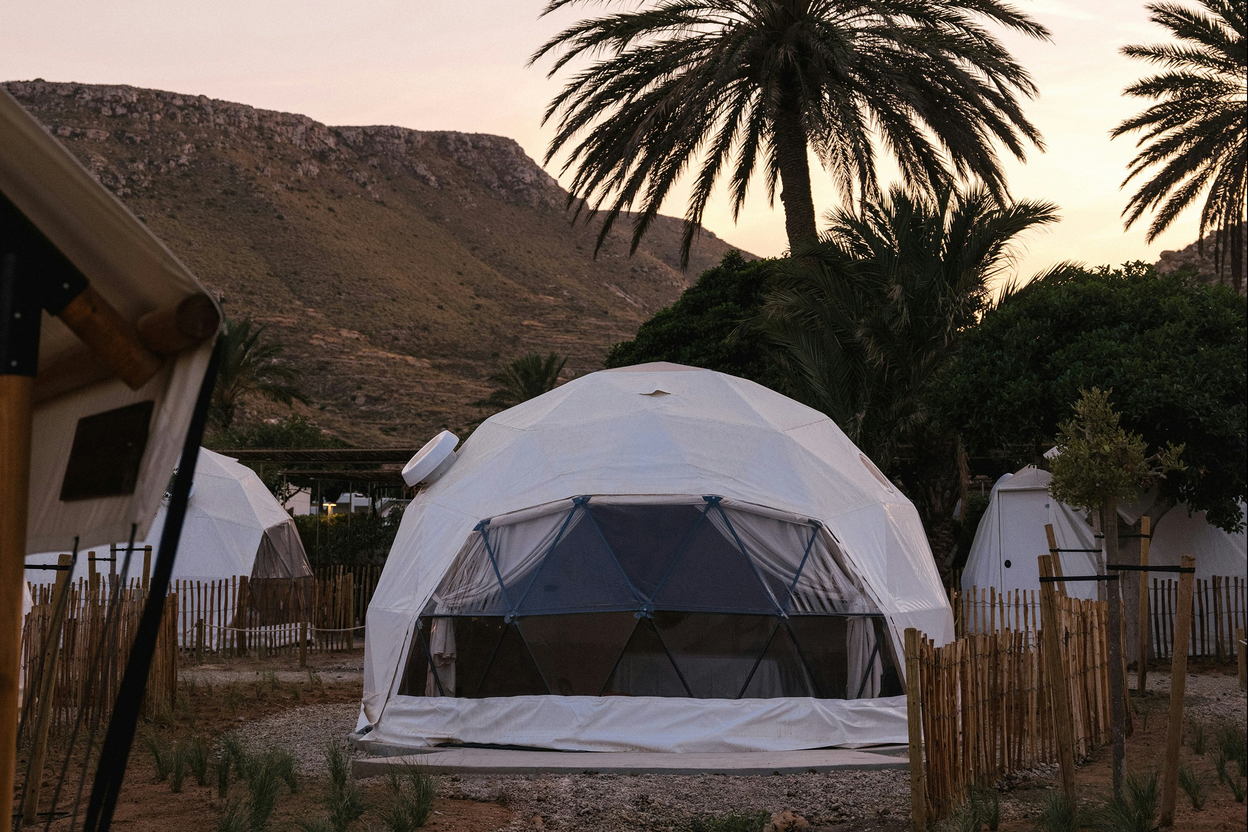 Glamping-Zelt mit geöffneter Front, daneben weitere Zelte, Palmen und Berge im Hintergrund bei Abendlicht.