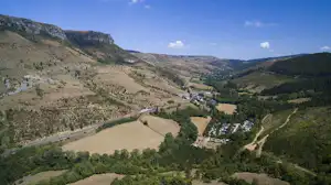 Panoramablick auf ein Tal mit Feldern, einem Fluss und einem Campingplatz am Hang zwischen grünen Hügeln und Bergen.