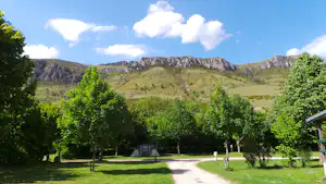 Campingwiese mit gepflastertem Weg, Bäumen und einem Wohnwagen im Hintergrund vor grünen Bergen.