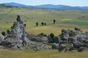 Felsformationen und ein kleines Dorf am Hang, dahinter weite grüne Hügel und Täler mit einzelnen Bäumen.