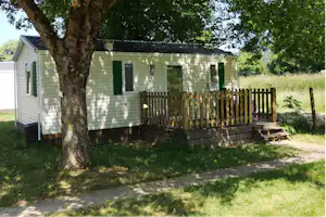 Mobilheim mit Holzterrasse am Baum, seitliche Fenster und Eingangstür, Rasenfläche und Weg am Campingplatz.