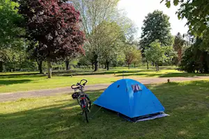 Blaues Campingzelt auf einer grünen Wiese, daneben ein Fahrrad am Weg, umgeben von Bäumen und Hecken.
