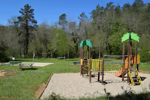 Spielplatz mit Klettergerüst und Rutsche auf gepflastertem Weg, grüne Wiese und Bäume im Hintergrund.