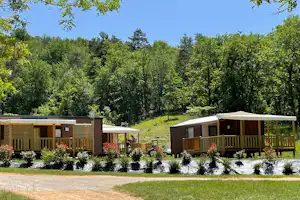 Camping-Bungalows mit Holzterrassen und Markisen, umgeben von Blumenbeeten und dichtem Wald im Hintergrund.