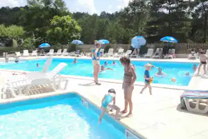 Schwimmbecken auf dem Campingplatz mit Kindern im Wasser, weißen Liegen und blauen Sonnenschirmen, umgeben von Bäumen.
