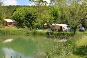 Campingplatz mit Wohnwagen und Zelt am Teich, grüner Rasen, Bäume und ein Auto am Stellplatz.