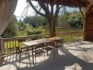 Überdachte Camping-Terrasse mit Holzdach, Tisch und Stühlen, Blick auf Bäume und einen See im Hintergrund.