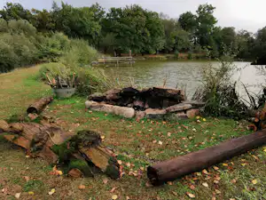 Feuerstelle mit Holzscheiten am Campingplatz am Fluss, Grasfläche und Bäume am Ufer.