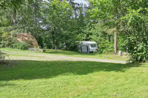 Grüner Campingplatz mit Rasenfläche, Bäumen und einem Wohnwagen am Rand des Weges.