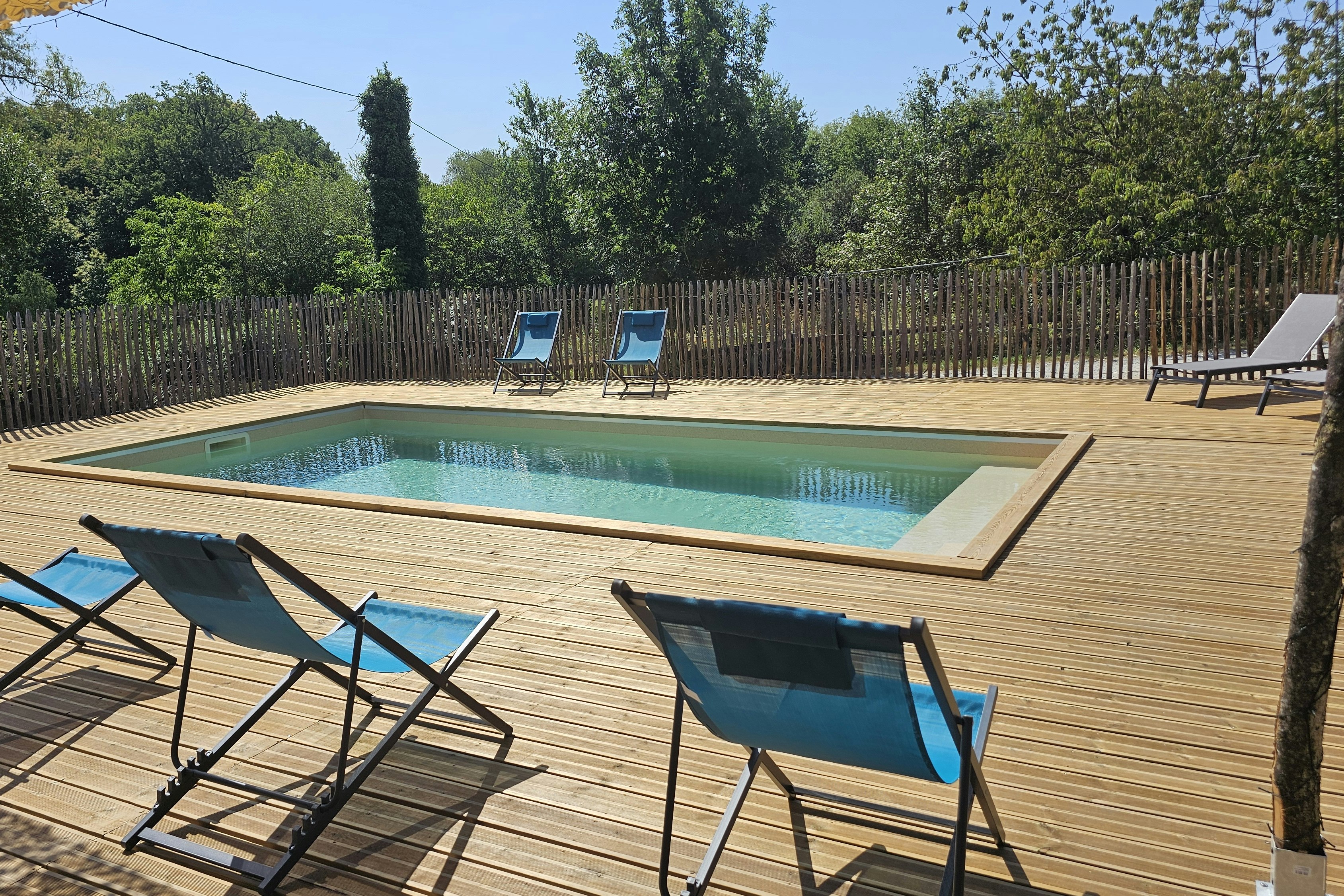 Schwimmbecken mit Holzdeck und zwei Liegestühlen am Pool, umgeben von Zaun und Bäumen im Campingbereich.