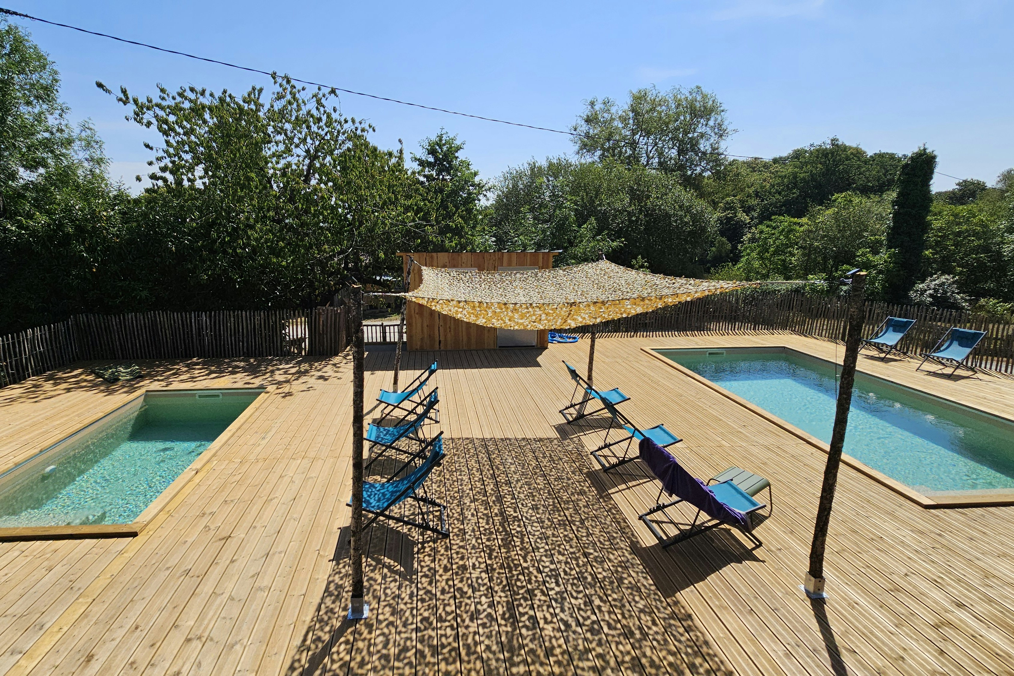 Zwei Holzterrassen mit zwei Swimmingpools, Liegestühlen und Sonnenschirm im Garten, umgeben von Hecken und Bäumen.