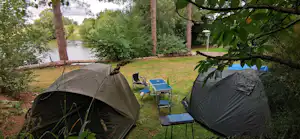 Zwei Campingzelte auf einem Rasenplatz am See, mit Campingstühlen und Tisch zwischen den Zelten, umgeben von Bäumen.