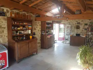 Innenraum eines Camping-Shops mit Holzregalen, Getränkeflaschen und Schränken, Steinwand und Fliesenboden, offene Tür zum Außenbereich.