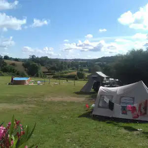 Zelte auf einem Campingplatz auf einer grünen Wiese, im Hintergrund Bäume und Hügel unter blauem Himmel.