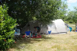 Zelte auf einem Campingplatz im Schatten von Bäumen, mit Sitzbereich und kleinen Tischen auf dem Rasen.