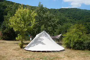 Weißes Campingzelt auf einer Wiese, umgeben von Büschen und Bäumen vor bewaldeten Hügeln.
