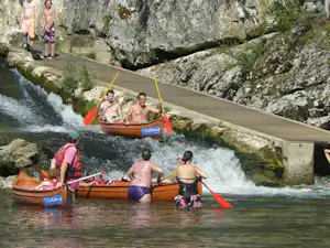 Rafting-Boote im Fluss mit Stromschnellen, Personen im Schlauchboot und auf dem Wasserweg zwischen Felsen und Bäumen.