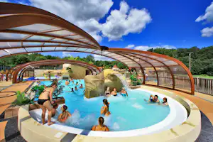 Überdachtes Camping-Schwimmbad mit mehreren Becken, Wasserspiel und Liegen, Menschen im Pool vor blauem Himmel.