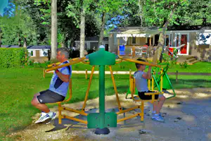 Zwei Kinder nutzen ein grünes Fitnessgerät im Campinggarten, im Hintergrund Bäume, Rasen und ein Gebäude mit Terrasse.