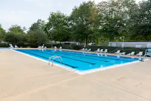 Schwimmbecken mit blauem Wasser, Leiter und Reihen weißer Liegestühle am Rand, dahinter Hecken und Bäume.