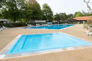 Schwimmbecken mit blauem Wasser, Liegestühlen am Rand und Poolbereich auf dem Campingplatz, umgeben von Bäumen und Gebäuden.