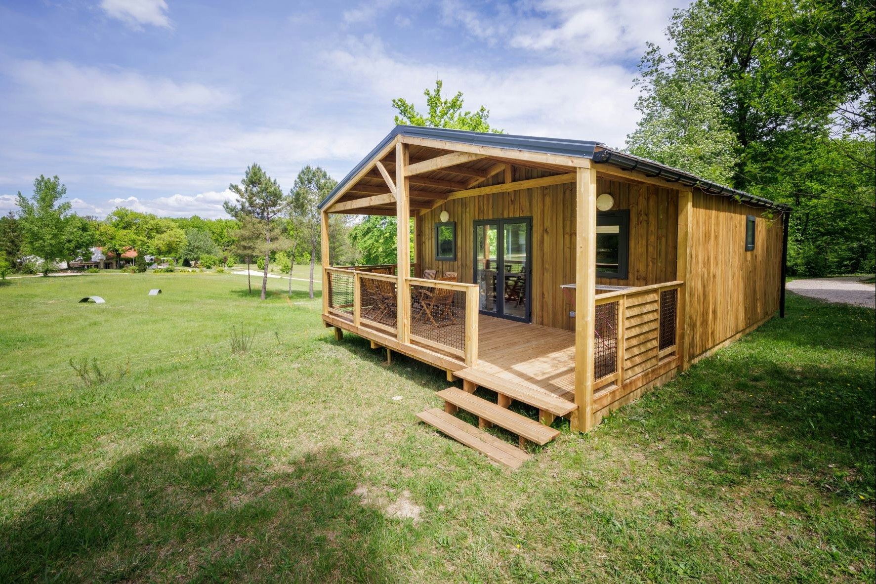 Holzhütte mit Veranda und Stufen auf einer grünen Wiese, umgeben von Bäumen und einem Weg.