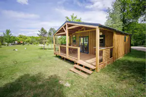 Holzhütte mit Veranda und Stufen auf einer grünen Wiese, umgeben von Bäumen und einem Weg.