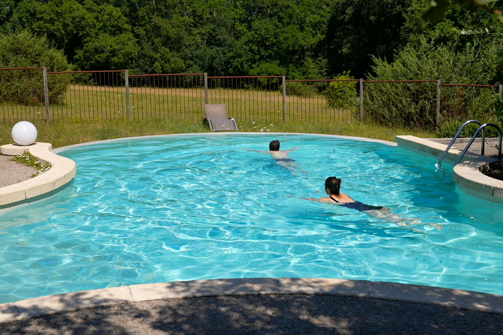 Campingpool mit türkisfarbenem Wasser, Einstiegstreppe und Liegestuhl im Grünen, umgeben von Zaun und Bäumen.