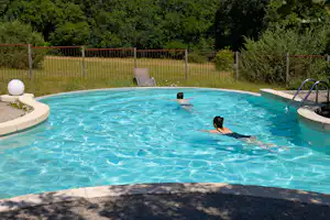 Campingpool mit türkisfarbenem Wasser, Einstiegstreppe und Liegestuhl im Grünen, umgeben von Zaun und Bäumen.