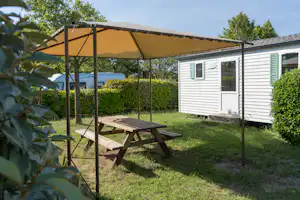 Stellplatz mit Wohnwagen und überdachter Terrasse, Holztisch und Bank im Garten mit Hecken und Bäumen.