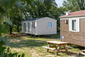 Weiße Wohnwagen-Mobilheim mit Vorhangfenster auf dem Campingplatz, daneben Picknicktisch und Rasen zwischen Bäumen.