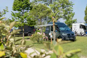 Wohnmobile auf einem Campingplatz im Grünen, zwischen Bäumen und Hecken, mit Stellplatznummer am Pfosten.