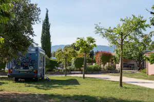 Wohnmobil auf einem Campingplatz mit Rasenfläche, Bäumen und gepflastertem Weg, im Hintergrund Gebäude und Hecken.