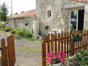 Steinbau mit Eingang, Holzzaun und Blumenbeeten am Weg; im Garten stehen Töpfe und ein großer Wassertank.