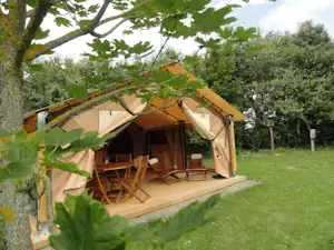 Großes Campingzelt auf Holzpodest mit Sitzgruppe und Tisch, umgeben von Rasen und Bäumen im Grünen.