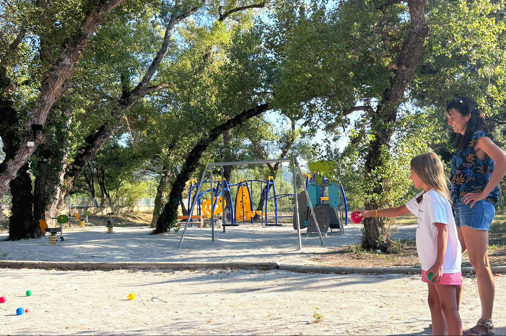 Spielplatz auf dem Campingplatz mit Schaukel und Rutsche im Schatten von Bäumen, daneben stehen ein Kind und ein Erwachsener.