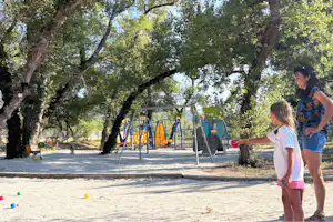 Spielplatz auf dem Campingplatz mit Schaukel und Rutsche im Schatten von Bäumen, daneben stehen ein Kind und ein Erwachsener.