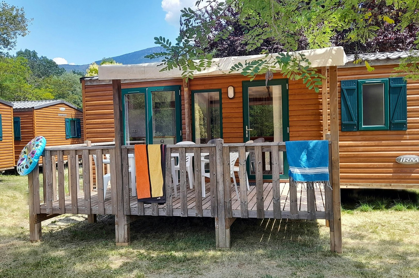 Wohnwagen mit Holzfassade und Veranda, davor Tisch und Stühle, im Hintergrund Bäume und Hügel.