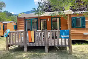 Wohnwagen mit Holzfassade und Veranda, davor Tisch und Stühle, im Hintergrund Bäume und Hügel.