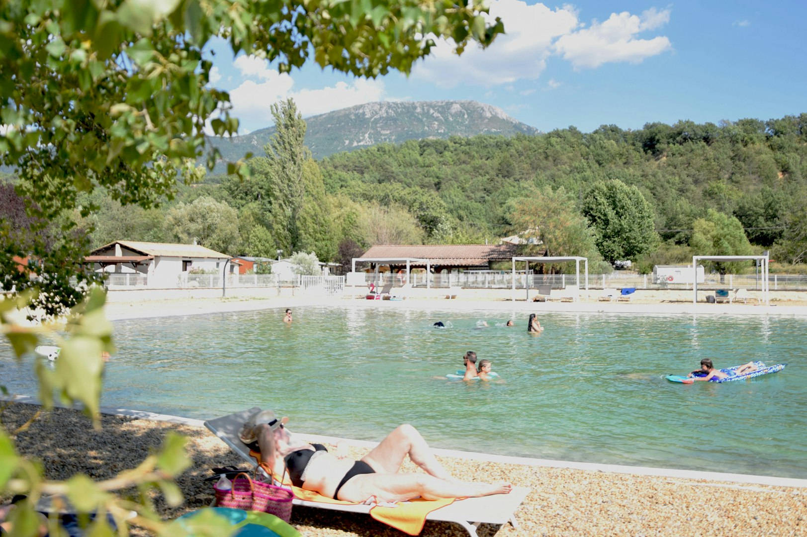 Schwimmbecken am Campingplatz mit türkisfarbenem Wasser, Liegen und Menschen im Hintergrund, umgeben von Bäumen und Gebäuden.