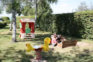Spielplatz auf dem Campingplatz: gelbe Federwippe, Sandkasten und Spielhaus im Grünen neben einem Holzkasten.