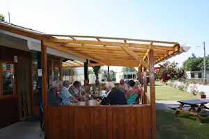 Holzpergola mit Tisch und Sitzbank, mehrere Personen beim Essen, daneben Rasen und Blumenbeete am Campingplatz.