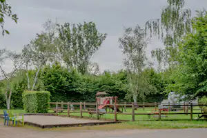 Spielplatz auf dem Campingplatz mit Rutsche und Sandfläche, umgeben von Hecken, Bäumen und Holzgeländer.