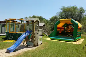 Spielplatz mit Rutsche und Hüpfburg auf dem Campingplatz, daneben Sandfläche, Zaun und Bäume im Hintergrund.
