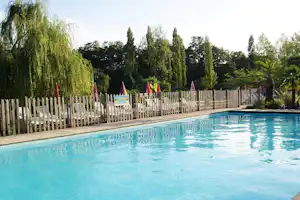 Schwimmbecken auf dem Campingplatz mit weißem Zaun, Bäumen und Palmen im Hintergrund.
