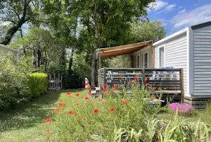 Wohnwagen mit Holzterrasse, Blumenbeet und Sitzbank im Garten, umgeben von Bäumen und Rasenfläche.