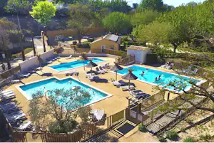 Camping-Poolanlage mit zwei Becken, Sonnenliegen und Sonnenschirmen, umgeben von Bäumen und kleinen Gebäuden.